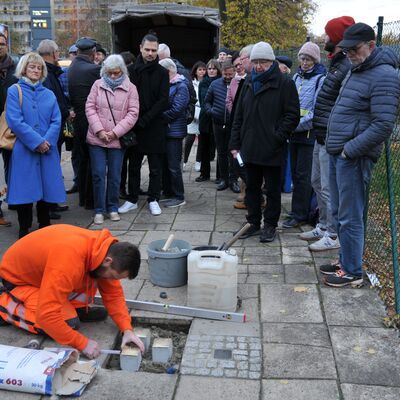 Foto: Ein Mann in oranger Arbeitskleidung verlegt Pflastersteine. Im Hintergrund eine Gruppe Menschen.