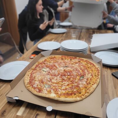 Bild vergr&ouml;&szlig;ern: Foto: Eine Pizza. Im Hintergrund Teller auf einem Tisch, an dem Kinder sitzen.