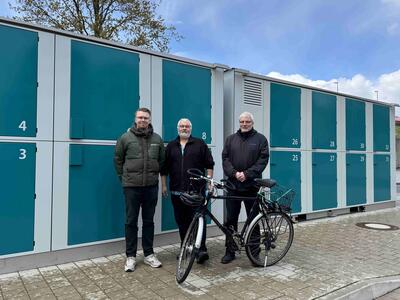 Bild vergr&ouml;&szlig;ern: Drei Männer stehen mit einem Fahrrad vor zwei großen Containern mit mehreren Türen und Nummern.