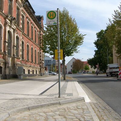 Eine neu sanierte Bushaltestelle. Links davon ein historisches Gebäude, rechts die Straße. Auf der gegenüberliegenden Straßenseite wird eine weitere Bushaltestelle saniert.