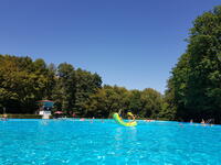 Bild vergr&ouml;&szlig;ern: Ein Freibad umgeben von Bäumen. Darüber blauer Himmel. Man sieht Menschen schwimmen und Kinder auf einem aufblasbaren Spielzeug sitzen.