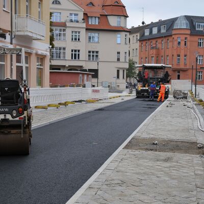 Eine Straßenbaustelle mit Baufahrzeugen und Bauarbeitern. Die Fahrbahn ist mit frischem, schwarzen Asphalt überzogen.