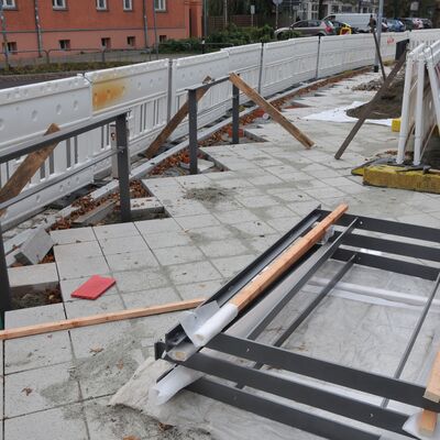 Eine Baustelle mit grauen Geländern, die zwischen Pflastersteinen eingebaut werden. Dahinter eine weiße Baustellen-Absperrung.