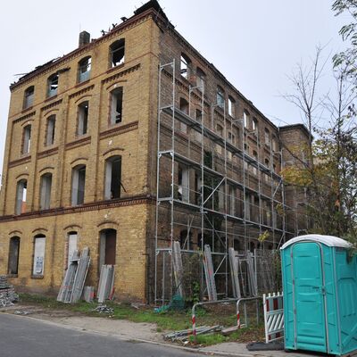 Ein altes Gebäude aus gelbem Backstein, das zum Teil eingefallen ist. Davor steht zum Teil ein Gerüst, rechts daneben ein türkises Dixi-Klo.