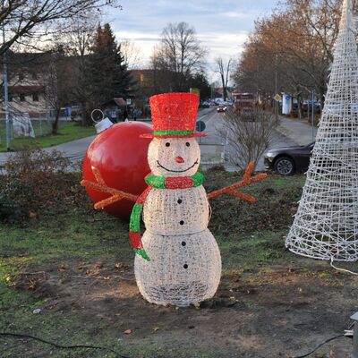 Ein Schneemann aus Plastik-Geflecht. Er ist weiß, hat braune Äste als Arme, trägt einen roten Hut und einen rot-grünen Schal, hat eine rote, karottenähnliche Nase und lächelt. Im Hintergrund eine große, rote Weihnachtskugel und ein großer weißer Kegel aus Plastikgeflecht. Im Hintergrund sieht man Autos und eine Straße.