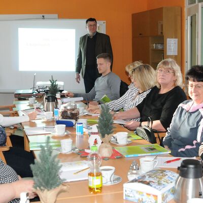 Foto: Mehrere Personen, sitzend an einem Tisch und stehend in einem Raum, schauen in Informationsbroschüren und sprechen darüber.