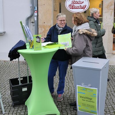 Foto: jeweils zwei Frauen im Gespräch, im Vordergrund ein Tisch mit Formularen und Broschüren sowie eine Einsteckurne und Informationsaufsteller
