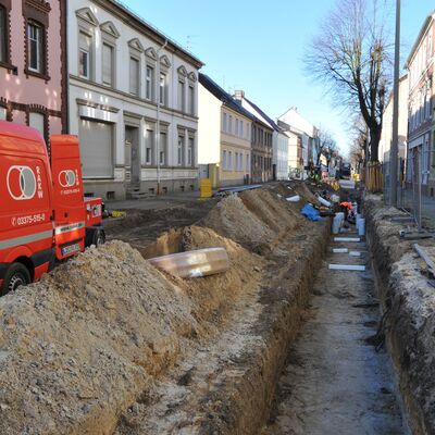 Foto: Eine Baustelle mit Graben, Fahrzeugen und Baumaterial zwischen zwei Häuserzeilen.