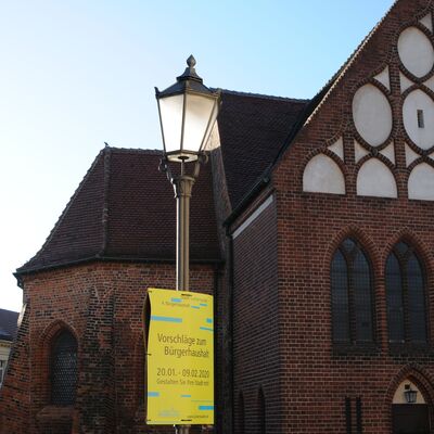 Foto: Plakat an einer Laterne im Vordergrund, dahinter ein historisches Kirchengebäude. Auf dem Plakat steht - Stadt Luckenwalde - vierter Bürgerhaushalt - Vorschläge zum Bürgerhaushalt - 20.01. bis 09.02.2020 - Gestalten Sie Ihre Stadt mit!- www.luckenwalde.de
