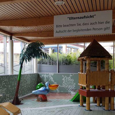 Foto: Kinderbereich in der Fläming-Therme. Ein Raum mit einer Holzdecke, gefliestem Boden und seichtem Wasser. Die Wände sind halb verglast und halb gefliest. An der Decke hängt ein Schild auf dem steht: "Elternaufsicht. Bitte beachten Sie, dass auch hier die Aufsicht der begleitenden Person gilt!" Darunter sieht man Spielgeräte und gelbe Liegen.