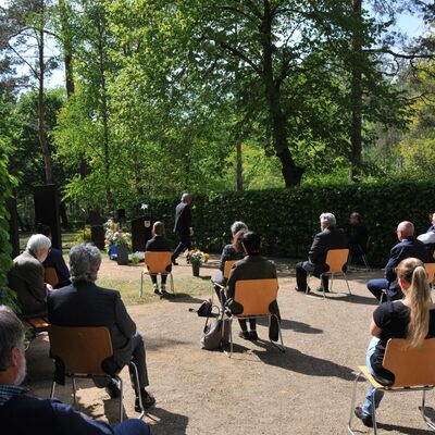 Foto: Männer und Frauen sitzen auf Stühlen, die im großen Abstand zueinander stehen. Vorne steht ein Mann bei Blumengestecken. Seitlich Hecken und viele Bäume.