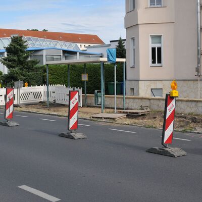 Foto: Baustelle an einer Bushaltestelle. Im Hintergrund Gebäude, vorne eine Straße mit Absperrbaken.