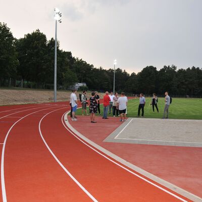 Foto: Eine Sportanlage mit roter Laufbahn. Eine Gruppe Menschen steht zwischen einer Sandgruppe und einem Fußballfeld.