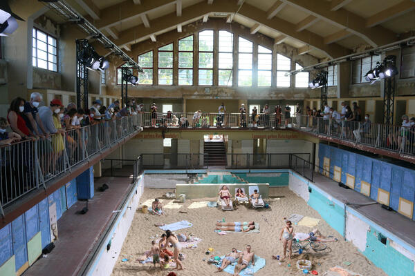 Bild vergr&ouml;&szlig;ern: Foto: Eine große Halle mit einem Schwimmbecken, das voll Sand gefüllt ist. Einige Menschen stehen am Rand, einige sitzen in Badeklamotten auf dem Sand.