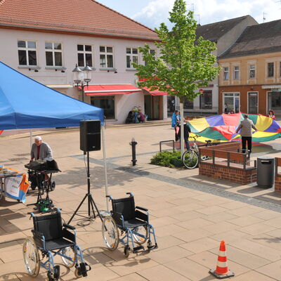 Foto: Ein Platz mit einem Stand und zwei Rollstühlen. Im Hintergrund Menschen mit einem großen, bunten Tuch.