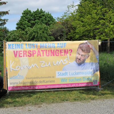 Foto: Zwei Männer befestigen ein buntes Banner im Grünen. Darauf ein Mann und der Schriftzug: Keine Lust mehr auf Verspätungen? Komm zu uns! Stadt Luckenwalde. Wir suchen Dich! www.luckenwalde.de/Karriere.