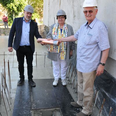 Foto: Zwei Männer und eine Frau mit Bauhelmen halten gemeinsam eine kupferne Röhre.