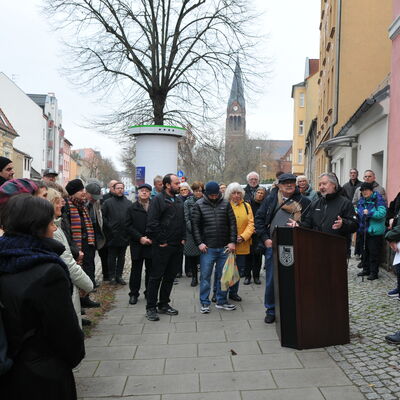 Foto: Eine Menge Menschen auf einem Gehweg vor einem Gebäude. In der Mitte stehen zwei Männer an einem Rednerpult.