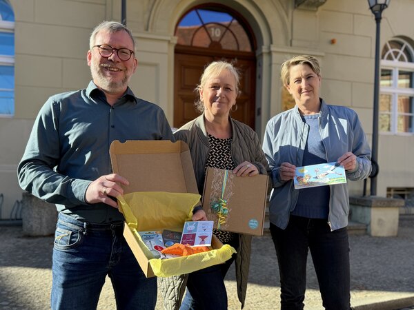 Bild vergr&ouml;&szlig;ern: Ein Mann und zwei Frauen stehen vor einem Gebäude und halten Präsente in der Hand