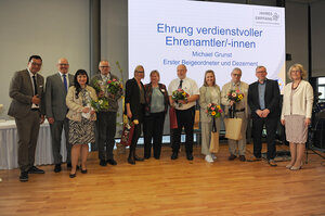Bild vergr&ouml;&szlig;ern: Foto: Eine Gruppe Menschen, zum Teil mit Blumensträußen in den Händen, vor einer Leinwand.