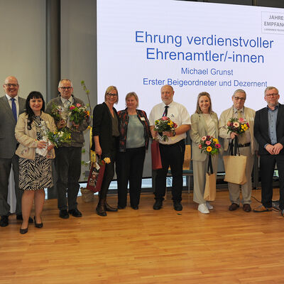 Foto: Eine Gruppe Menschen, zum Teil mit Blumensträußen in den Händen, vor einer Leinwand.