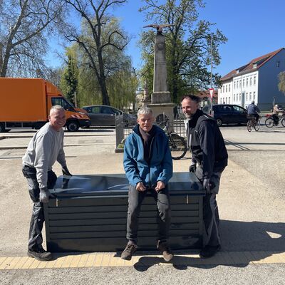 Foto: Ein Mann sitzt auf einer grauen, großen Kiste. Zwei weitere Männer stehen links und rechts daneben.