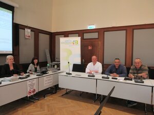 Zwei Frauen und drei Männer sitzen in einem Raum an Tischen und schauen in die Fotokamera.