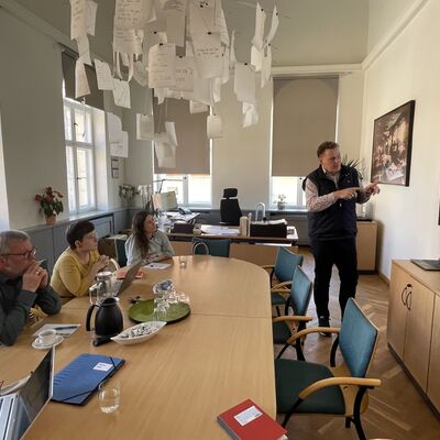 Foto: Ein Mann steht vor einem Bildschirm. An einem Tisch sitzen zwei Frauen und ein Mann.