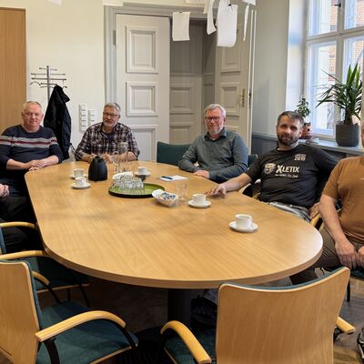 Sechs Männer sitzen um einen Besprechungstisch und lächeln in die Kamera.