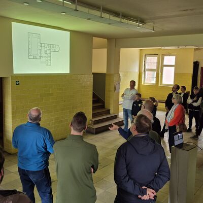 Bild vergr&ouml;&szlig;ern: Foto: Menschen stehen in einem Gebäude. Ein Gebäudeplan wird auf eine Wand projiziert.