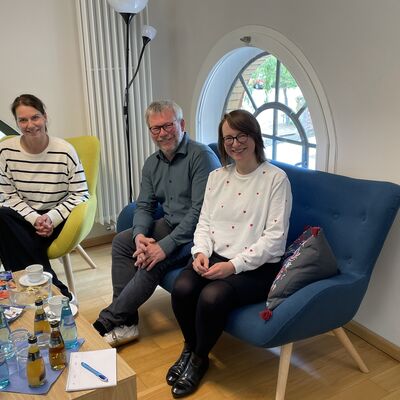 Foto: Zwei Frauen und ein Mann sitzen auf farbigen Sesseln zusammen um einen Tisch.