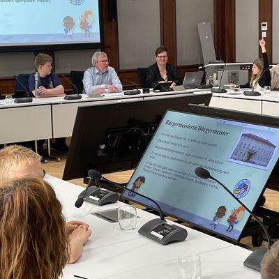 Foto: Erwachsene und Kinder sitzen in einem großen Raum mit einer Leinwand und Monitoren.