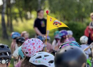 Bild vergr&ouml;&szlig;ern: In der Mitte des Bildes ist eine Fahrradwimpel im Fokus. Rundherum sieht man Kinderköpfe mit Fahrradhelmen