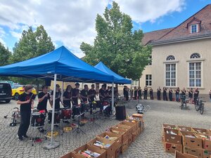 Bild vergr&ouml;&szlig;ern: Foto: Zahlreiche Musiker unter blauen Pavillons vor einem großen Gebäude. Davor stehen Kartons mit Büchern.