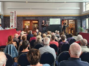 Bild vergr&ouml;&szlig;ern: Foto: Viele Menschen sitzen in einer großen Halle, vorne stehen zwei Menschen auf einer kleinen Bühne.