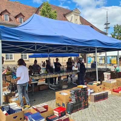 Bild vergr&ouml;&szlig;ern: Foto: Menschen und zahllose Kartons mit Büchern unter blauen Pavillons.