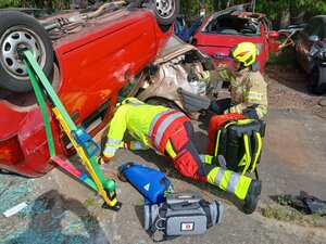 Bild vergr&ouml;&szlig;ern: Foto: Zwei Menschen in Einsatzklamotten vor einem roten Auto, das auf dem Dach liegt.