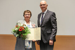 Bild vergr&ouml;&szlig;ern: Foto: Ein Mann steht neben einer Frau, die einen Blumenstrauß und eine Urkunde hält.