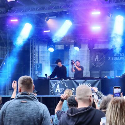 Bild vergr&ouml;&szlig;ern: Ein DJ legt auf einer Bühne auf und wird dabei von einem Kameramann gefilmt. Menschen im Publikum schauen zur Bühne.