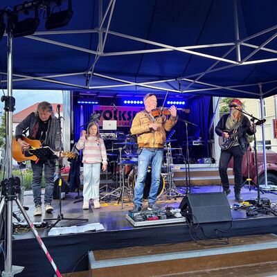 Bild vergr&ouml;&szlig;ern: Eine Band mit männlichen Musikern und ein junges Mädchen stehen auf einer Bühne unter einem Dach.