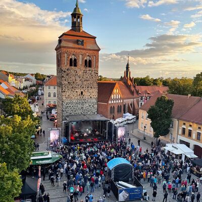Bild vergr&ouml;&szlig;ern: Blick auf einen gefüllten Marktplatz, auf dem eine Bühne und ein großer Turm dahinter stehen.