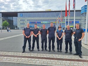 Bild vergr&ouml;&szlig;ern: Foto: Eine Gruppe Menschen in Einsatzklamotten steht vor einer Messehalle.