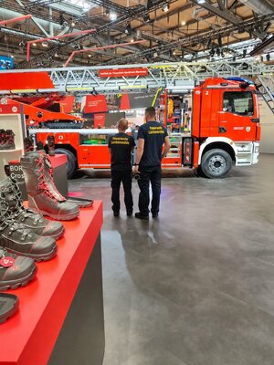 Bild vergr&ouml;&szlig;ern: Foto: Zwei Männer in Einsatzklamotten stehen vor einem Feuerwehrfahrzeug. Hinter ihnen stehen Stiefel auf einem roten Regal.