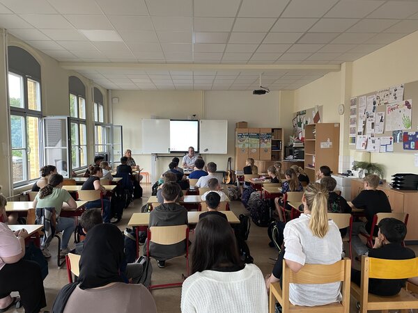 Foto: Ein Klassenzimmer voller Kinder. Vorne in der Mitte sitzt ein Mann vor einer digitalen Tafel.