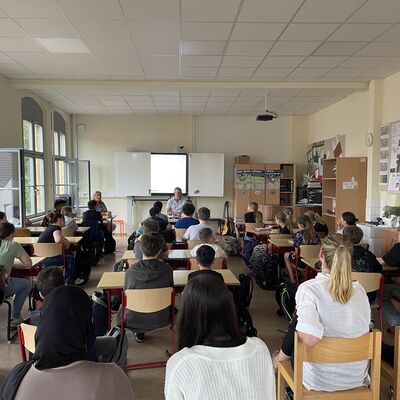Foto: Ein Klassenzimmer voller Kinder. Vorne in der Mitte sitzt ein Mann vor einer digitalen Tafel.