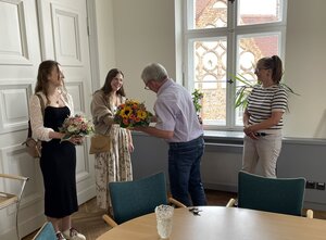 Bild vergr&ouml;&szlig;ern: Foto: Ein Mann übergibt einer jungen Frau einen Blumenstrauß. Daneben stehen zwei weitere Frauen, von denen eine auch einen Blumenstrauß hält.