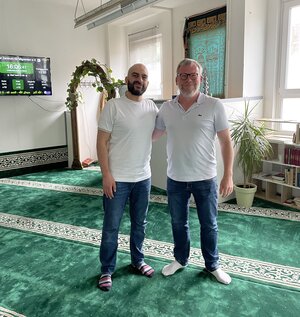 Bild vergr&ouml;&szlig;ern: Foto: Zwei Männer stehen in einem Raum mit grünem Teppich.