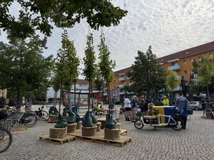 Bild vergr&ouml;&szlig;ern: Bäume stehen auf Paletten. Hinter ihnen sind Menschen bei einer Veranstaltung.