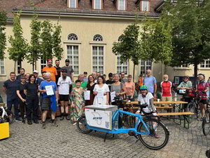 Mehrere Menschen stehen gemeinsam unter Bäumen vor einem Gebäude. Im Vordergrund befindet sich ein Fahrrad.