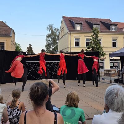 Foto: Menschen beobachten ein Theaterstück mit fünf rotgekleideten Personen auf Stelzen.